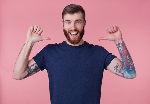 Portrait of young happy attractive red-bearded young guy , wearing a blue t-shirt, broadly smiling, enjoy himself, with arms raised up and pointing fingers at himself isolated over pink background.