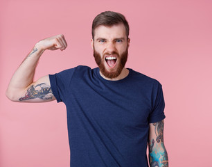 Young screaming red-bearded handsome manful guy demonstrates biceps and strength, looking at the...
