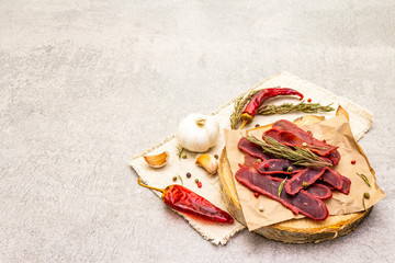 Basturma, dried tenderloin of beef meat, jerky, thinly sliced. Dry rosemary, pepper mix, chili, garlic on vintage linen cloth. Delicious food on a wooden board, stone background, copy space.