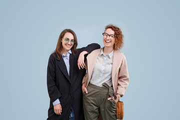 Portrait of laughing girls with hands on shoulders and glasses. Young happy girlfriends over blue isolated background. Smiling young female in casual coat having fun. Look at camera and each other.