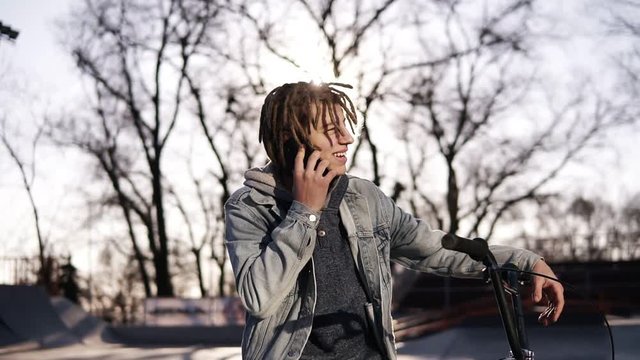 Portrait of happy handsome teenage boy with dreadlocks, using mobile phone while riding a BMX bike, smiling, messaging friends via social networks. People, lifestyle and communication concept