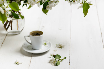 Spring still life. Cup of coffee with apple tree flowers and petals of flowers are on white cafe table outside in blooming garden