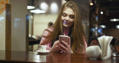 Young woman in the rose sweater holding a smartphone in hands and typing on its screen while chatting in the living room