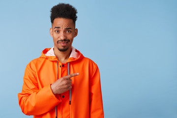 Unhappy young african american guy in orange rain coat looks at the camera inquiringly wants to draw your attention, points fingers at copy space on right. Stands over blue background.