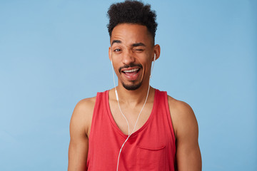 Close up of young african american guy in a great mood,wears in a red jersey, listens cool music, winks and smiles broadly stands over blue background.