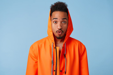 Portrait of amazed young african american dark skinned guy in orange rain coat, heard that his favorite band is releasing a new album, amazed by the news, says lips wow. Stands over blue background.