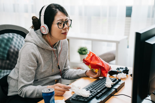 Upset Lazy Asian Woman Nerd Crying Looking At Pc Computer Screen Watching Sad Movie With Headphone. Unhappy Girl Geek Eat Snack Chip Junk Food In Hand Wipes Away Tears With Used Tissue On Table.