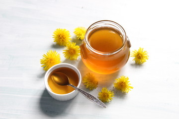 Dandelion jam in glass pot, bowl with spoon and yellow flowers on white background