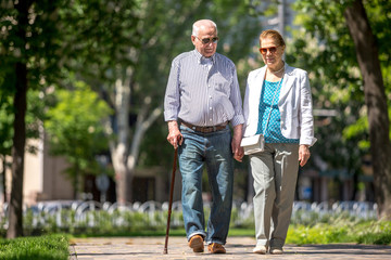 Cheerful senior couple having good time in city park, walking, laughing and enjoying sunny day. Old people wearing color clothes ans sunglasses