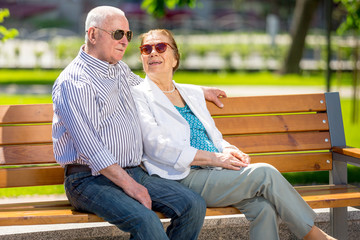 Fototapeta premium Cheerful senior couple having good time in city park, walking, laughing and enjoying sunny day. Old people wearing color clothes ans sunglasses