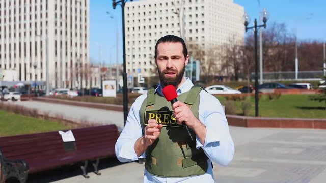 Medium shot, male television journalist in a bulletproof jacket talking to a microphone in front of skyscrapers in the financial district of the city.