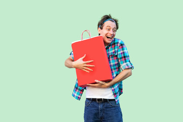 It is my! Crazy happy young shopoholic man in white t-shirt and checkered shirt standing, hugging shopping bags with toothy smile and opened mouth. Studio shot, green background, isolated,indoor