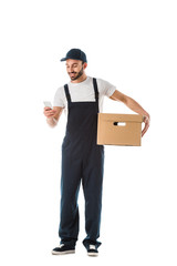 smiling delivery man using smartphone while holding cardboard box isolated on white