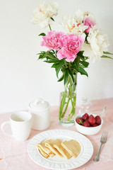 pancakes with strawberries and coffee on the table near a vase with peonies on a white background