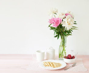 pancakes with strawberries and coffee on the table near a vase with peonies on a white background