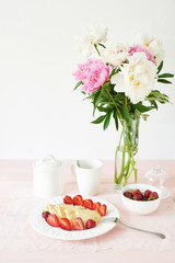 pancakes with strawberries and coffee on the table near a vase with peonies on a white background