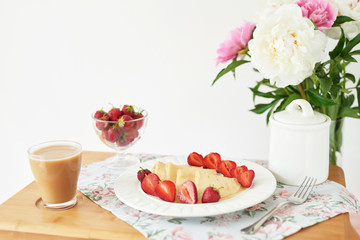 pancakes with strawberries and coffee on the table near a vase with peonies on a white background