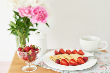 pancakes with strawberries and coffee on the table near a vase with peonies on a white background