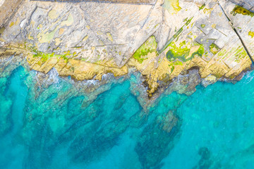 Aerial top view of sea waves hitting rocks on the rocky beach with green algae with turquoise sea water. Seascape in the coastline, drone shot.