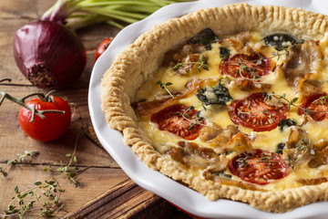 Kish open pie with tomatoes, bacon, spinach in white baking dish on wooden background