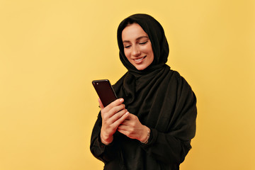 Lovely happy smiling arabian woman wearing black burqa looking on smartphone over isolated yellow background