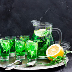 Summer cold green drink with tarragon, mint and lemon in glass decanter and small shots with ice, one in focus in focus. Sparkling beverage, alcohol drink on dark background. Square with copy space