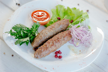 Meat kebabs on white plate with green herbs and red sauce.
