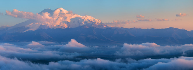 Fototapeta premium Beautiful sunset in the Caucasus Mountains, view towards Elbrus from the Bermamyt plateau. Snow-capped peaks above the clouds. Tourism in Russia.