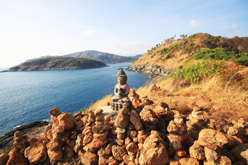 Beautiful Pyramid shape of stones arranged with Buddha statue in zen on rock mountain in seascape of sunset and sea horizon with Dry grass field on Phrom Thep Cape in Phuket island, Thailand.