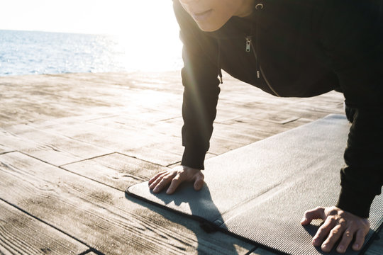 Photo of strong sportsman 20s in tracksuit lying on fitness mat and doing plank exercise by seaside