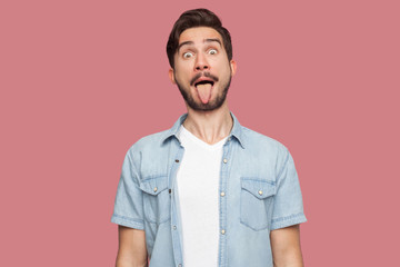 Fototapeta premium Portrait of funny crazy handsome bearded young man in blue casual style shirt standing with big eyes, tongue out and looking at camera. indoor studio shot, isolated on pink background.