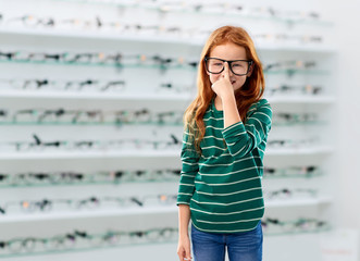 education, vision and childhood concept - smiling red haired student girl in glasses and green striped shirt over optics store background