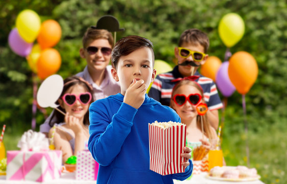 Fast Food, Childhood And People Concept - Boy In Blue Hoodie Eating Popcorn From Striped Paper Bucket Over Group Of Friends At Birthday Party In Summer Park Background