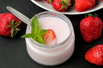 delicious strawberry yogurt in a jar and fresh ripe strawberries and a mint leaf on a black concrete table.