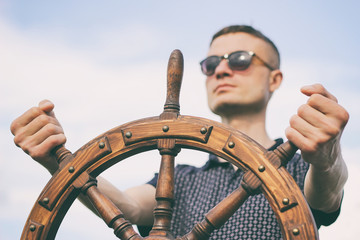 A man controls the yacht, hands on the steering wheel, handwheel on sky background