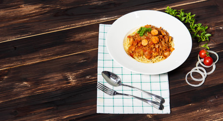 Spaghetti sausage tomato sauce in white plate on top with parsley and beside with spoon, fork, tomato, onion slices and green leaf all in dark wood background, left copy space
