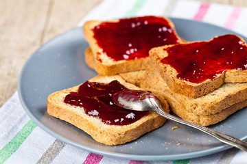 Fresh toasted cereal bread slices with homemade cherry jam and spoon on grey plate closeup on grey...