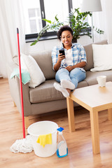 household and technology concept - happy african american woman or housewife with smartphone resting after home cleaning