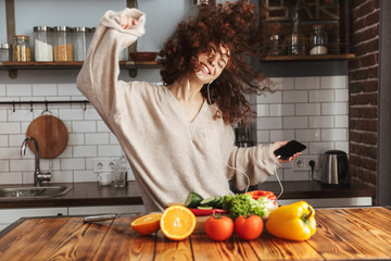 Image of young beautiful woman listening to music on mobile phone while cooking at home