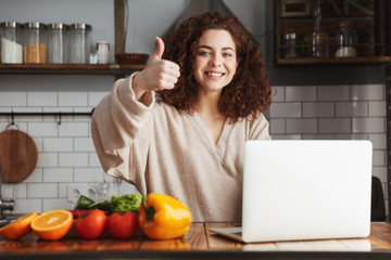 Image of young caucasian woman using laptop while cooking in kitchen interior at home