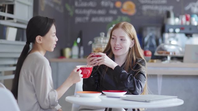 Medium shot of two young female colleagues enjoying talking over coffee in cafe. Asian and Caucasian businesswomen sitting at table and chatting