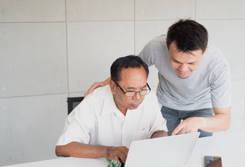 Young asian man in casual shirt with feeling confidence teach senior asian man with feeling confuse to use computer notebook in modern office background, family activity, copy space on top left