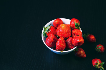 White bowl with fresh red strawberries on a dark wooden table close up. Spring or summer backgruond. Healthy food ingredients theme concept.