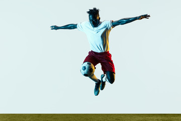 Naklejka na ściany i meble Young african-american male football or soccer player in sportwear and boots kicking ball for the goal in neon light isolated on white background. Concept of healthy lifestyle, professional sport