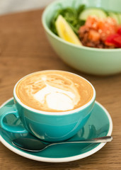 A cup of cappuccino with trendy latte art on a saucer with a coffee spoon. In the background a healthy breakfast bowl. Healthy eating concept, morning concept.