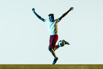 Fototapeta na wymiar Young african-american male football or soccer player in sportwear and boots kicking ball for the goal in neon light isolated on white background. Concept of healthy lifestyle, professional sport