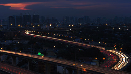 Fototapeta na wymiar Motion blur of car light in city highway at dawn. Cross intersection of urban highways. Background have silhouette of cityand sunset purple orange sky.