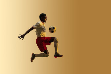 Fototapeta na wymiar Young african-american male football or soccer player in sportwear and boots kicking ball for the goal in jump in neon light on gradient background. Concept of healthy lifestyle, professional sport.