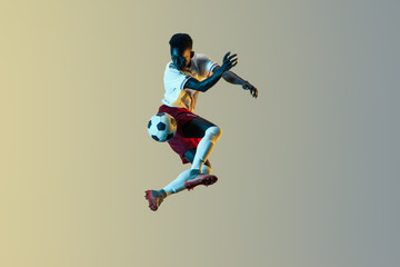 Fototapeta na wymiar Young african-american male football or soccer player in sportwear and boots kicking ball for the goal in jump in neon light on gradient background. Concept of healthy lifestyle, professional sport.