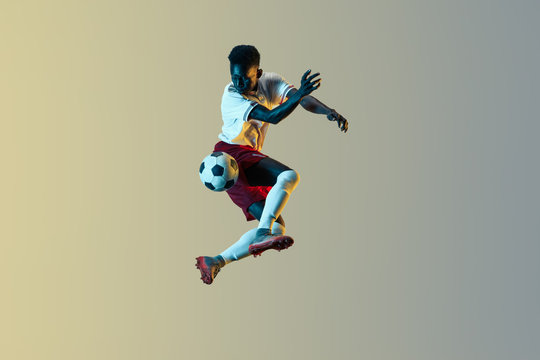 Young african-american male football or soccer player in sportwear and boots kicking ball for the goal in jump in neon light on gradient background. Concept of healthy lifestyle, professional sport. - Powered by Adobe
